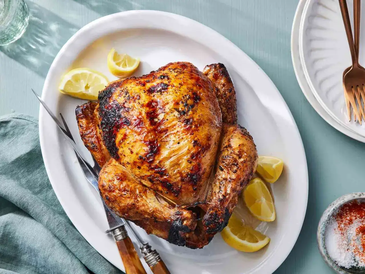 Un appétissant poulet à la broche doré et croustillant, servi sur un plat blanc avec des quartiers de citron. Des couverts à découper sont prêts.