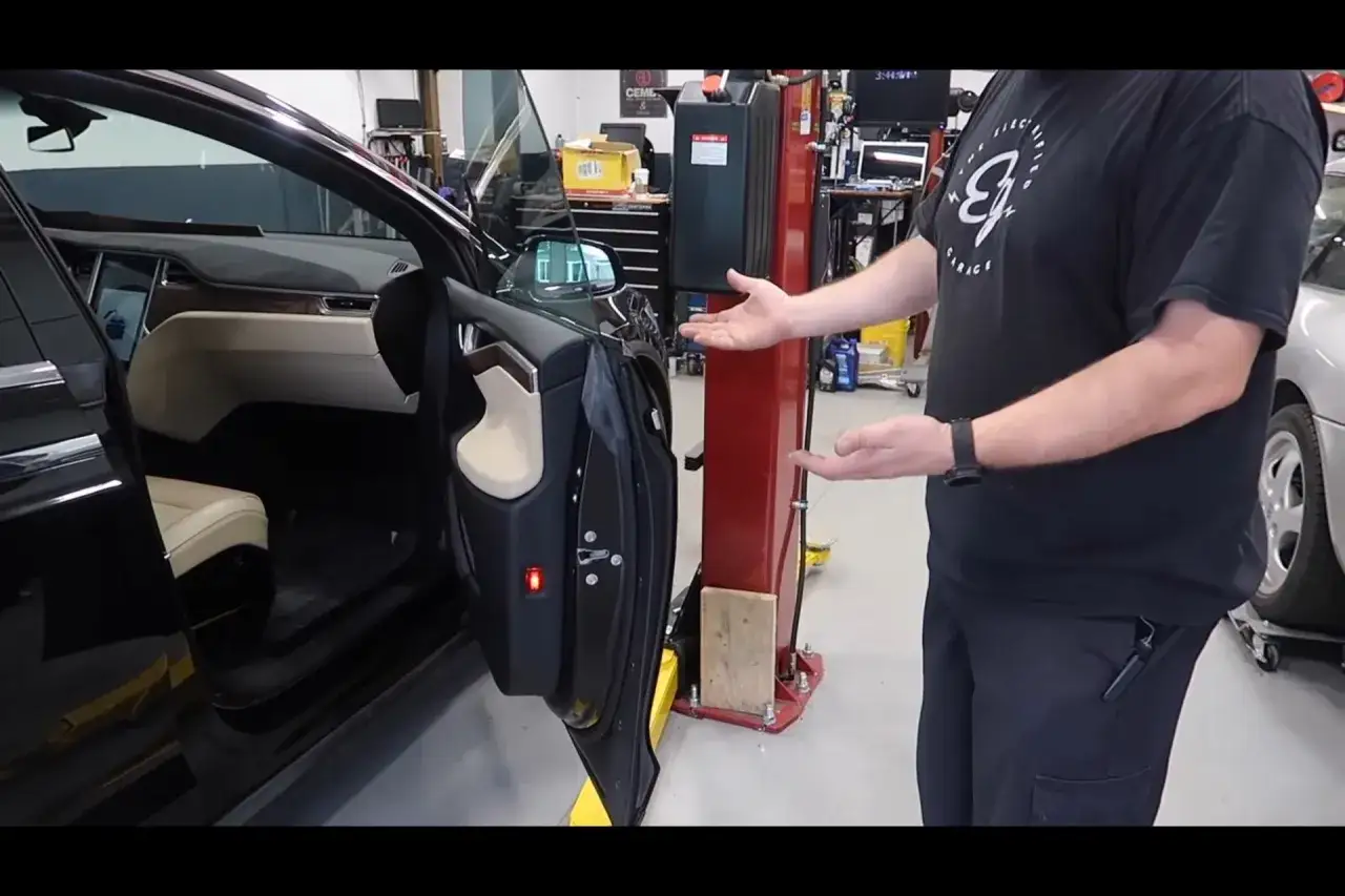 Wnętrze czarnego Tesla X z otwartymi drzwiami, jasną tapicerką i ekranem. Mężczyzna w czarnej koszulce pokazuje coś obok.