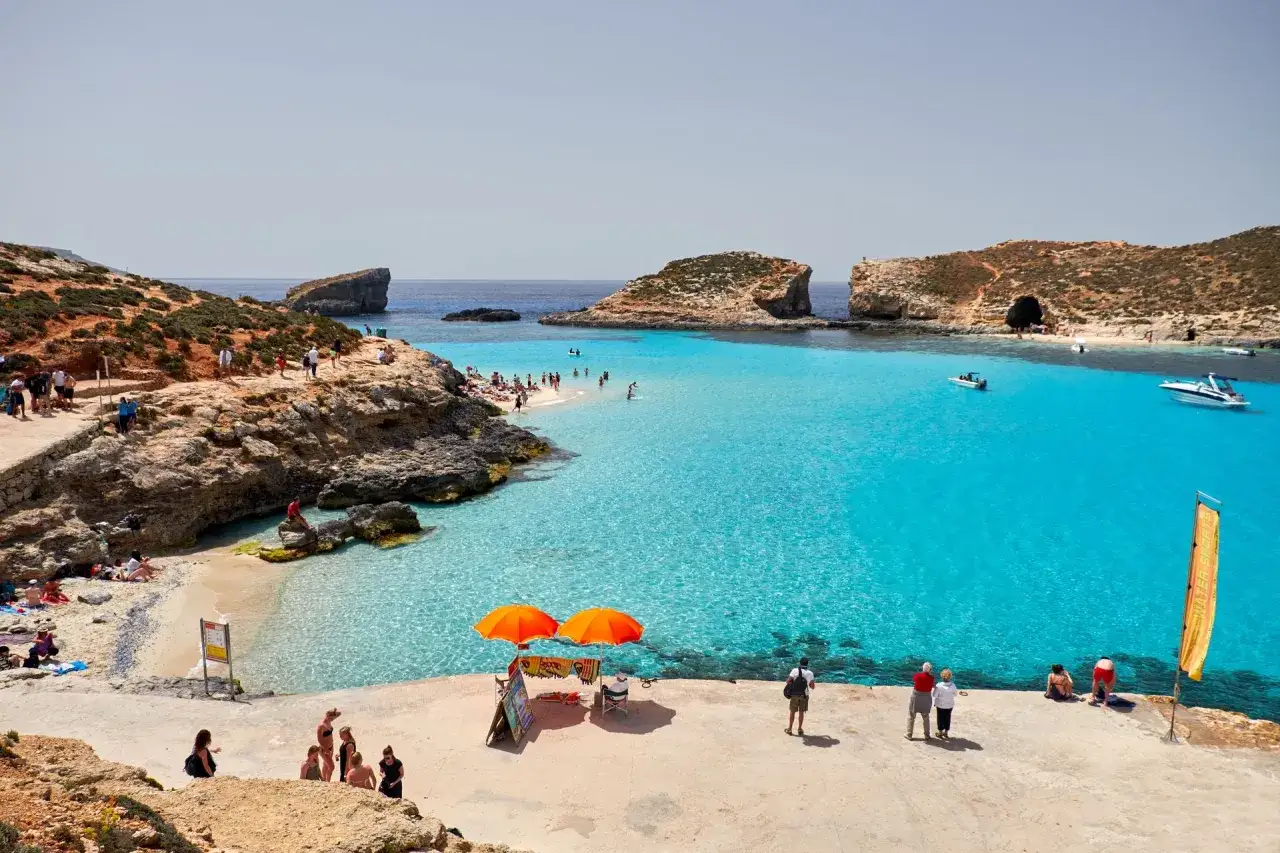 Malta ceny: Błękitna Laguna przyciąga turystów. Słoneczny dzień, turkusowa woda, plaża i łodzie. Idealne miejsce na wakacje.