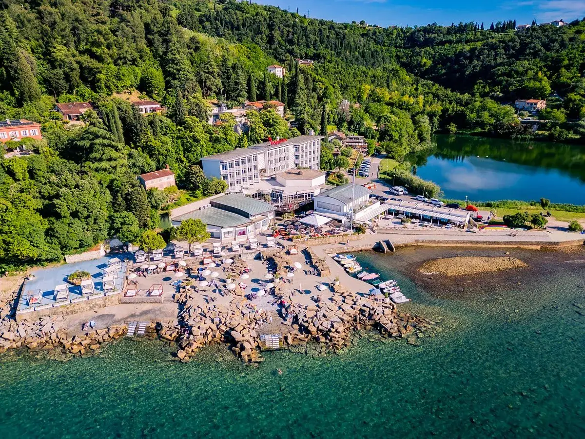 Słowenia morze: malownicza plaża z leżakami, restauracja i hotel na tle zielonych wzgórz. W oddali spokojna zatoka i łodzie.