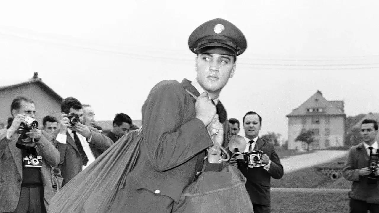 Elvis Presley im Armeedienst, umgeben von Paparazzi. Er blickt nachdenklich, w&auml;hrend Fotografen mit Kameras auf ihn richten.