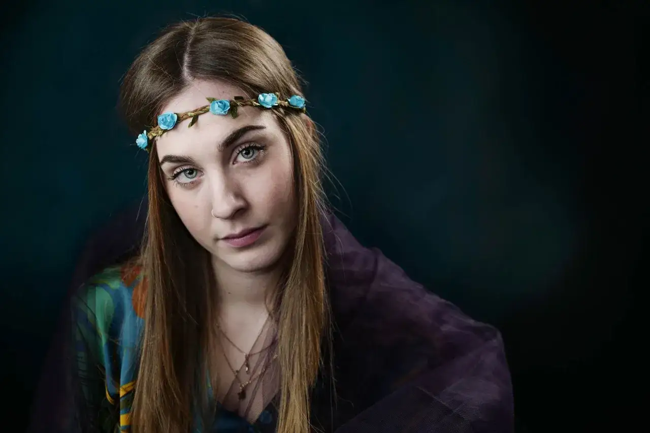 Giovane donna con coroncina di fiori azzurri e velo viola, i suoi occhi chiari catturano la luce in suggestivi schemi di luce.