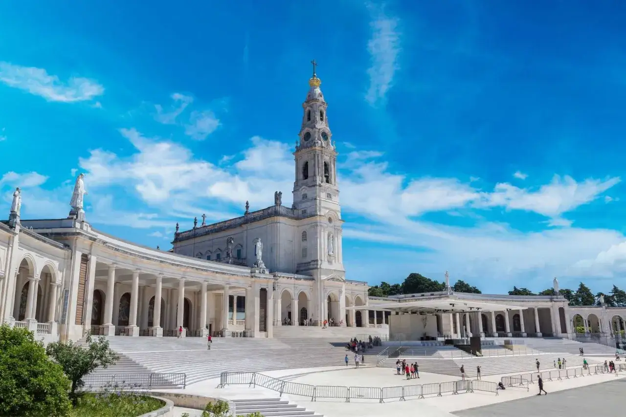 The Sanctuary of Fatima, Portugal, stands majestically under a bright blue sky with scattered clouds. Pilgrims gather on the grand staircase leading to the basilica.