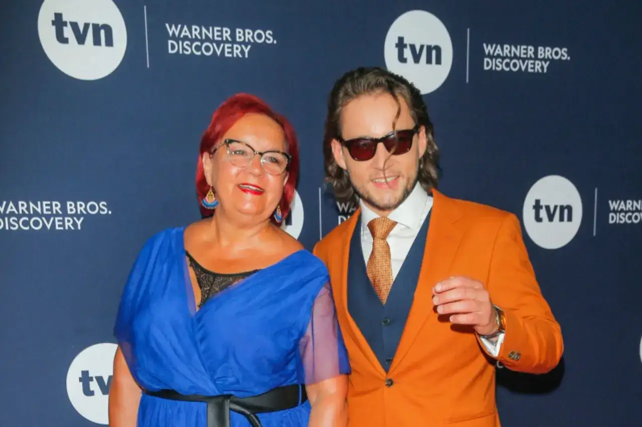 Joachim Zeiske w pomarańczowej marynarce i jego towarzyszka w niebieskiej sukni pozują na tle logo TVN i Warner Bros. Discovery.