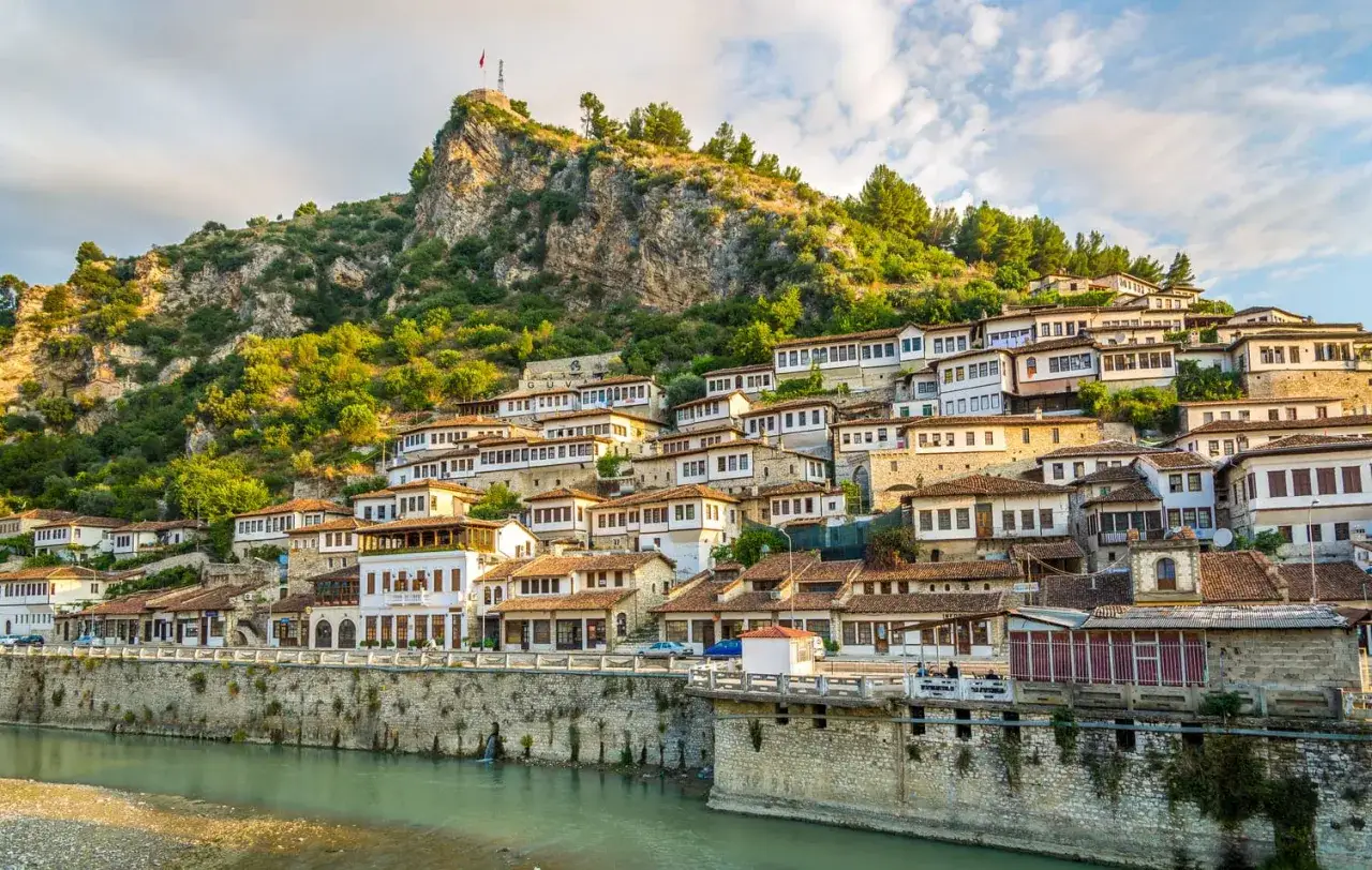Vue d'ensemble du paysage albanais : maisons blanches aux toits de tuiles rouges s'étageant sur une colline verdoyante, dominée par une forteresse.
