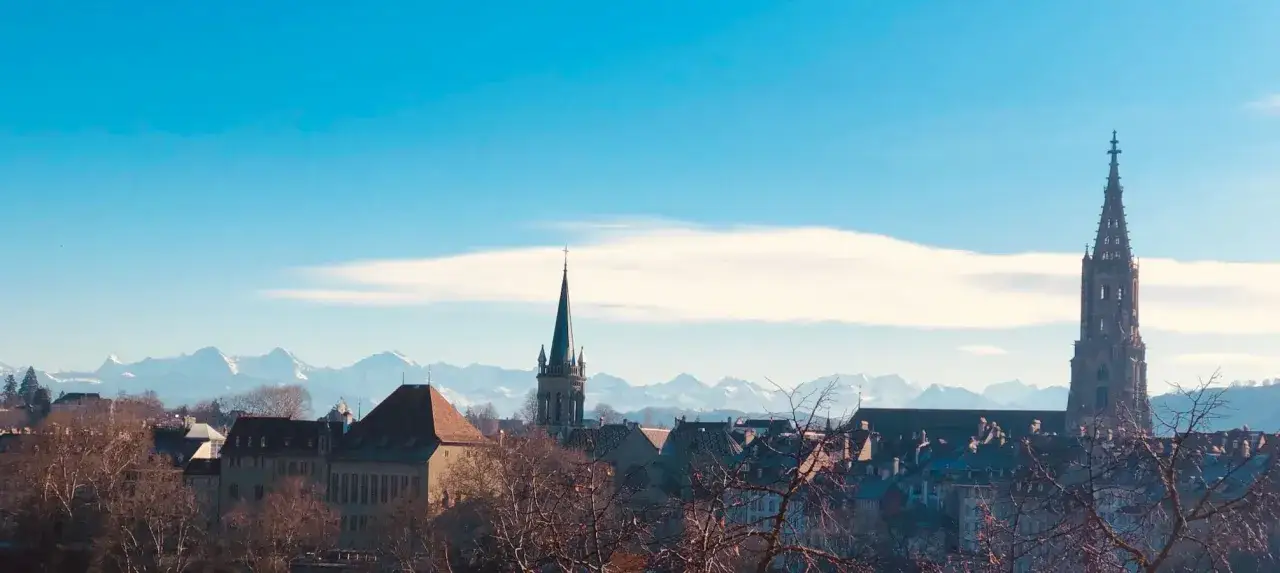 Blick von einer Besucherplattform in Bern auf die Alpen. Die T&uuml;rme der M&uuml;nsterkirche und eine weitere Kirche ragen &uuml;ber die D&auml;cher der Stadt.