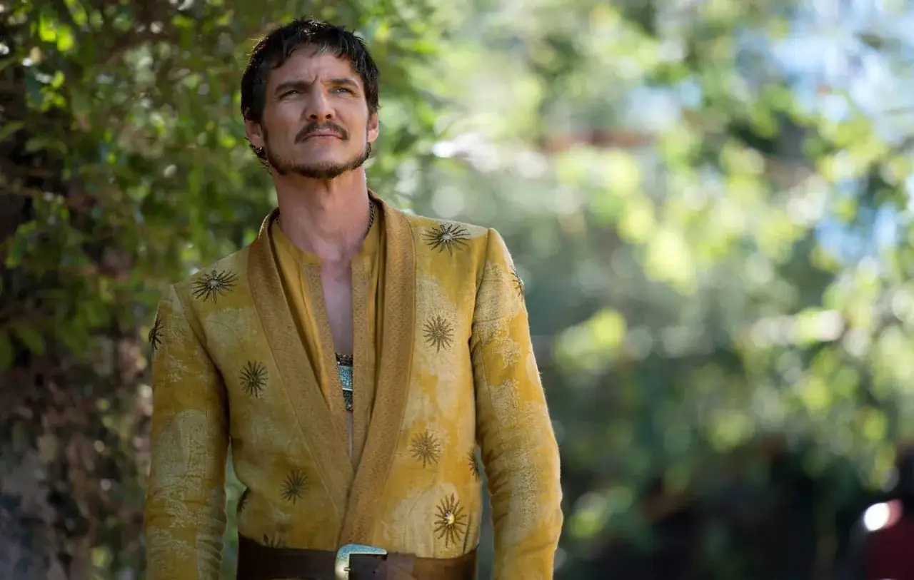 Oberyn Martell, książę Dorne, w swojej złotej zbroi z haftowanymi słońcami, patrzy w górę. Jego charakterystyczna broda i wąsy dodają mu uroku. To scena z serialu 