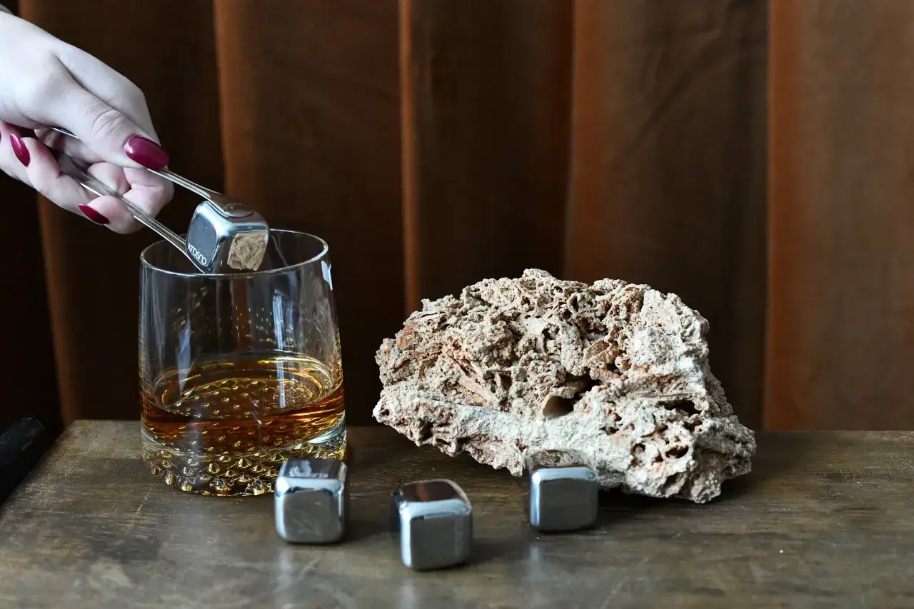 Dłoń z czerwonymi paznokciami wkłada metalowy kostkę do szklanki z whisky. Czy whisky się chłodzi? Obok leżą kolejne kostki i kamień.