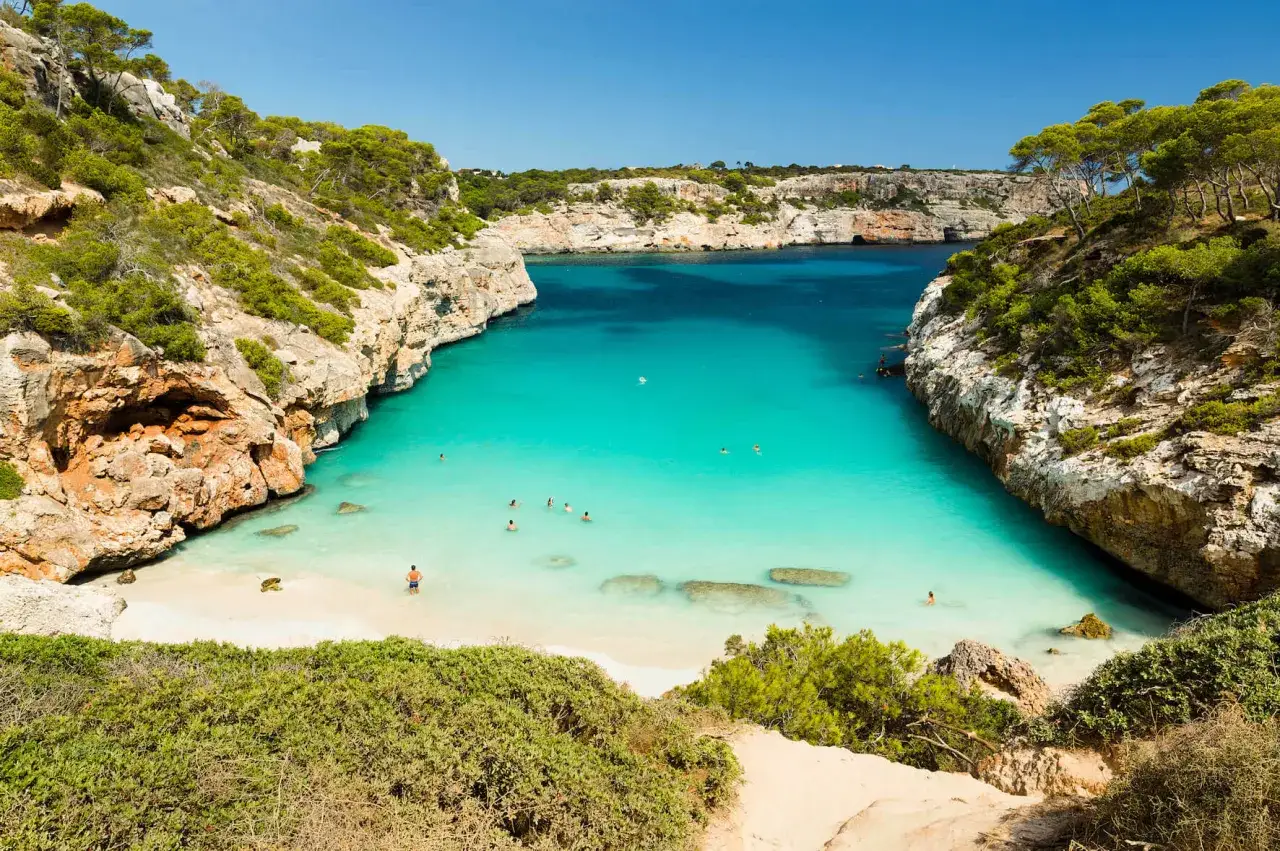 Bahía de aguas turquesas y cristalinas, rodeada de acantilados rocosos y vegetación exuberante. Un paraíso de **playas blancas** donde nadan personas.