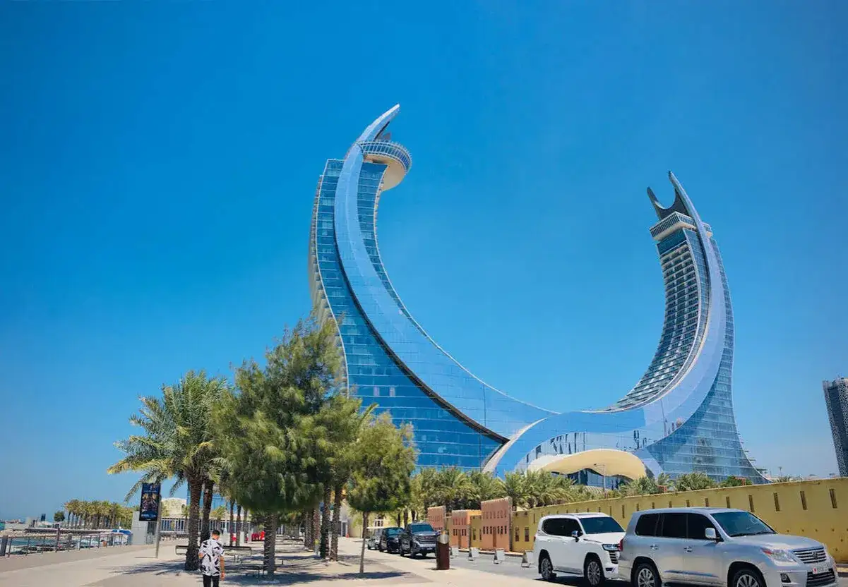 Moderne Architektur in Katar: Zwei geschwungene Glasgebäude ragen in den blauen Himmel. Palmen säumen die Promenade, auf der Autos parken.