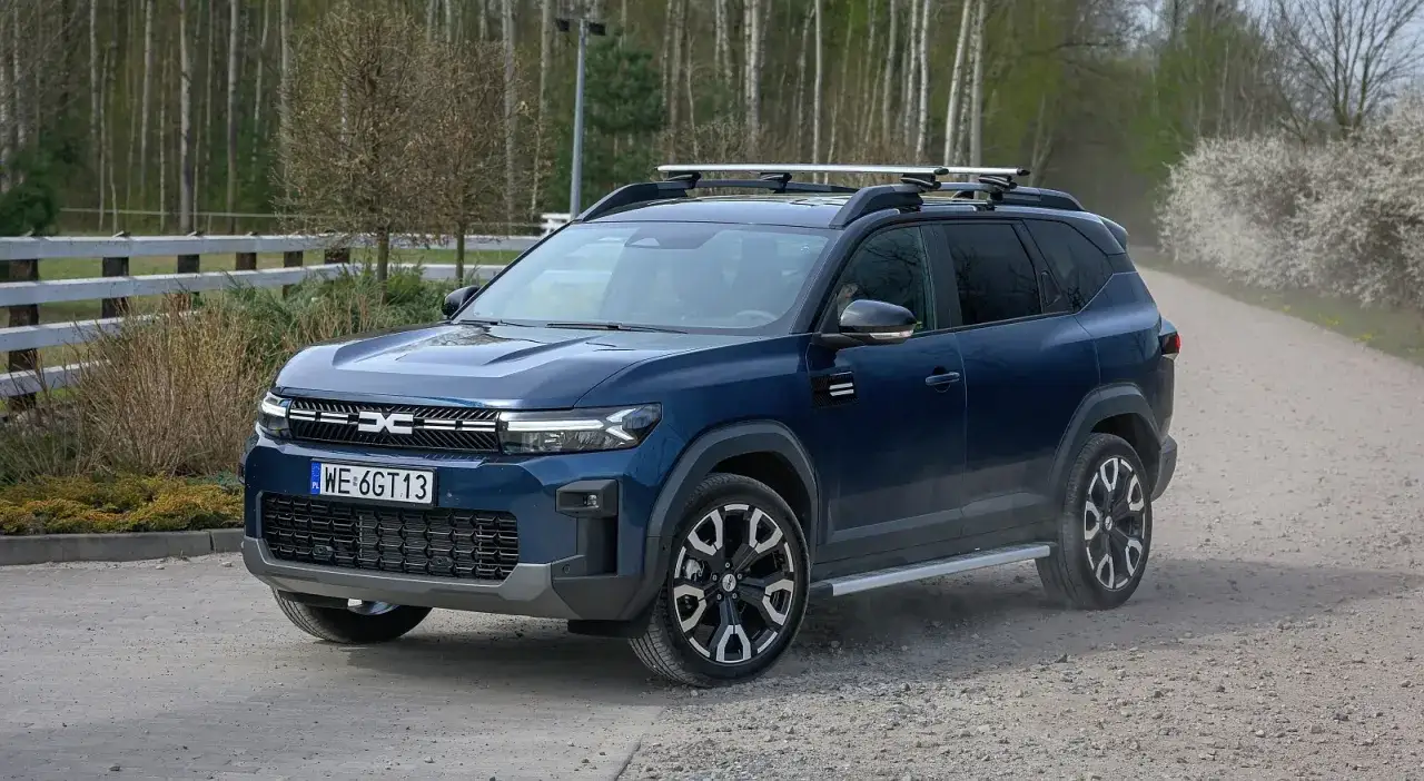Ciemnoniebieski SUV z relingami dachowymi, jeden z samochodów z fabrycznym LPG, jedzie po szutrowej drodze.