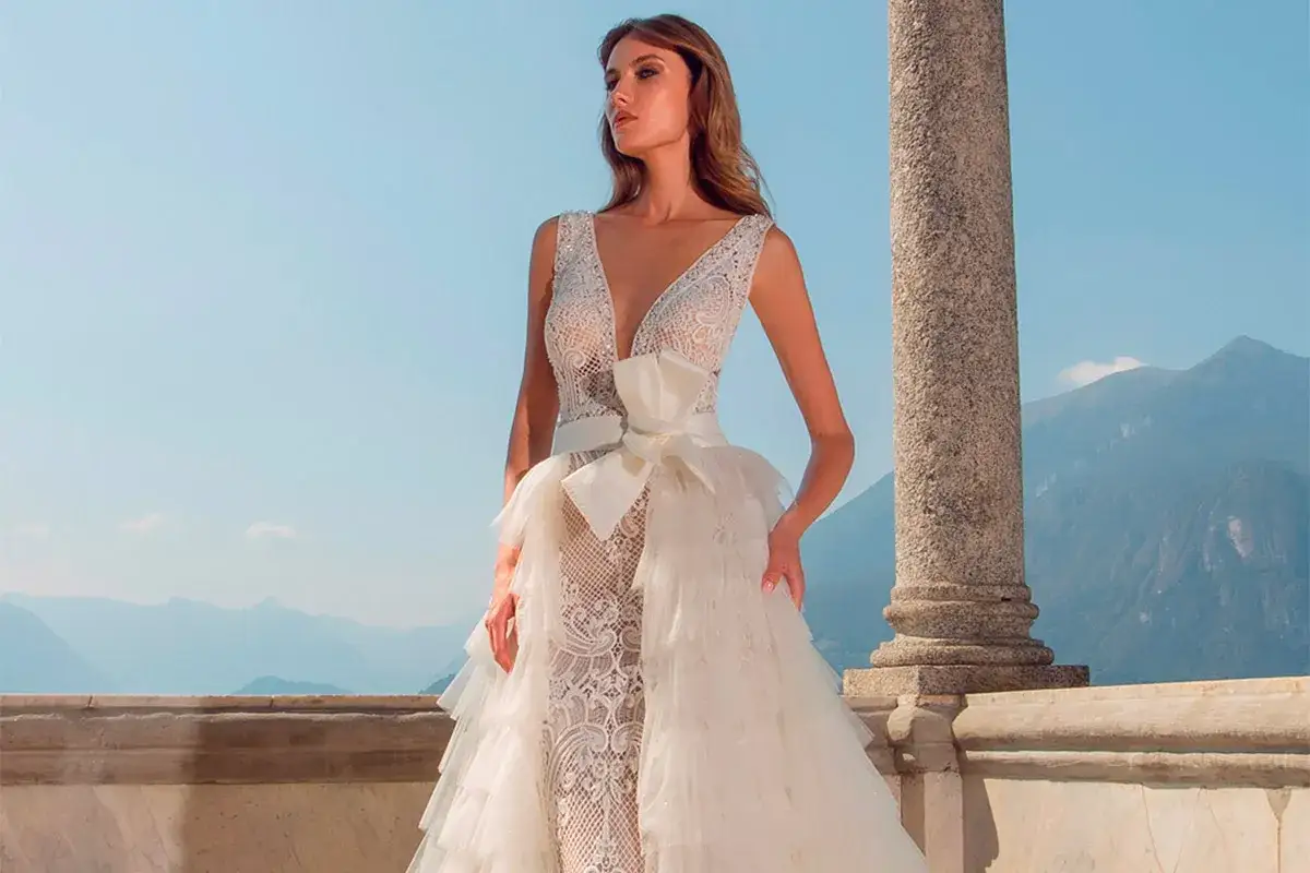 Modella con abiti da sposa riutilizzabili in pizzo e tulle, con fiocco in vita, su un balcone con vista sulle montagne.