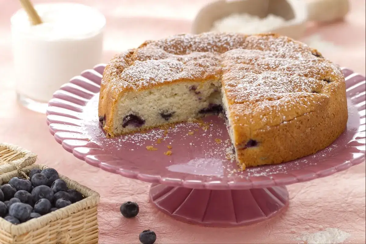 D&eacute;licieux g&acirc;teau sans oeuf aux myrtilles, saupoudr&eacute; de sucre glace, servi sur un plat rose. Un panier de myrtilles fra&icirc;ches est &agrave; c&ocirc;t&eacute;.