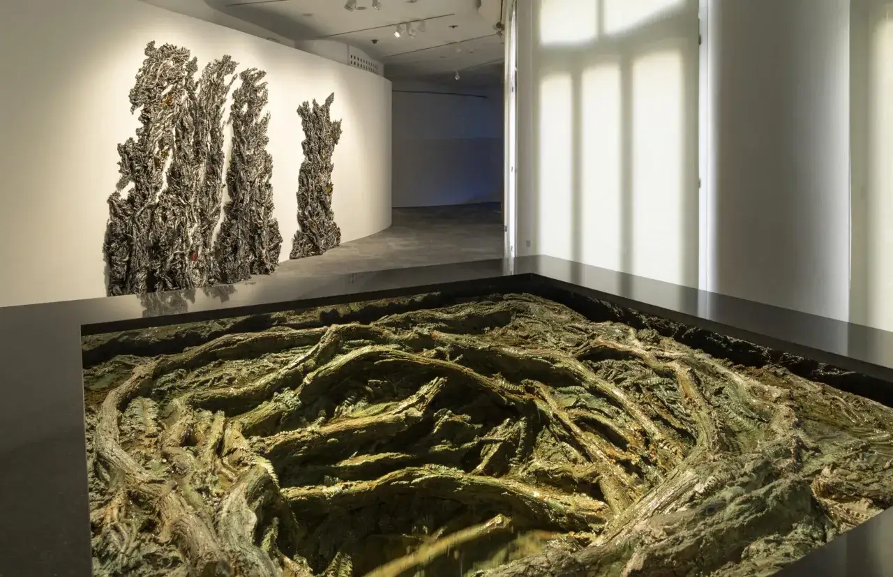 Instalación de Cristina Iglesias: esculturas oscuras y texturizadas en la pared, y un intrincado paisaje de raíces verdes y marrones en un foso.