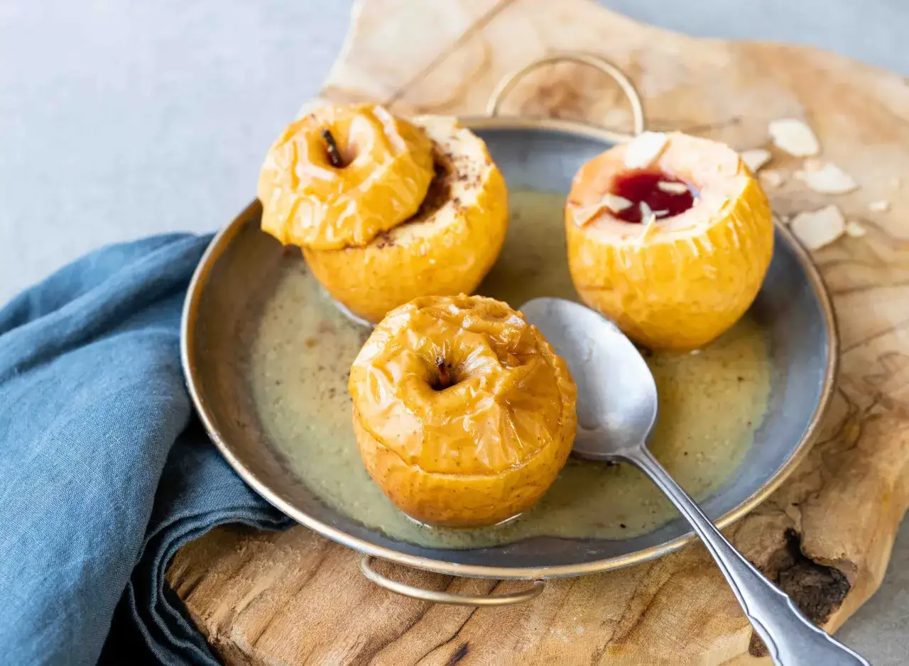 Trois pommes au four dorées et caramélisées, garnies de cannelle et de confiture, baignant dans un jus sucré sur un plateau métallique.