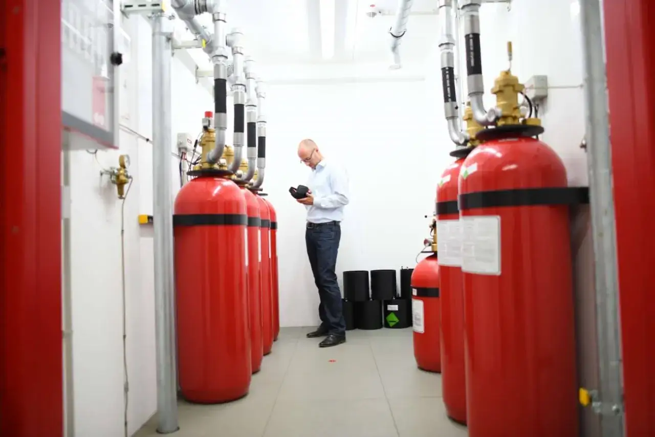 Mann pr&uuml;ft Gasflaschen f&uuml;r den Serverraum Brandschutz. Rote Zylinder mit Rohren und Ventilen.