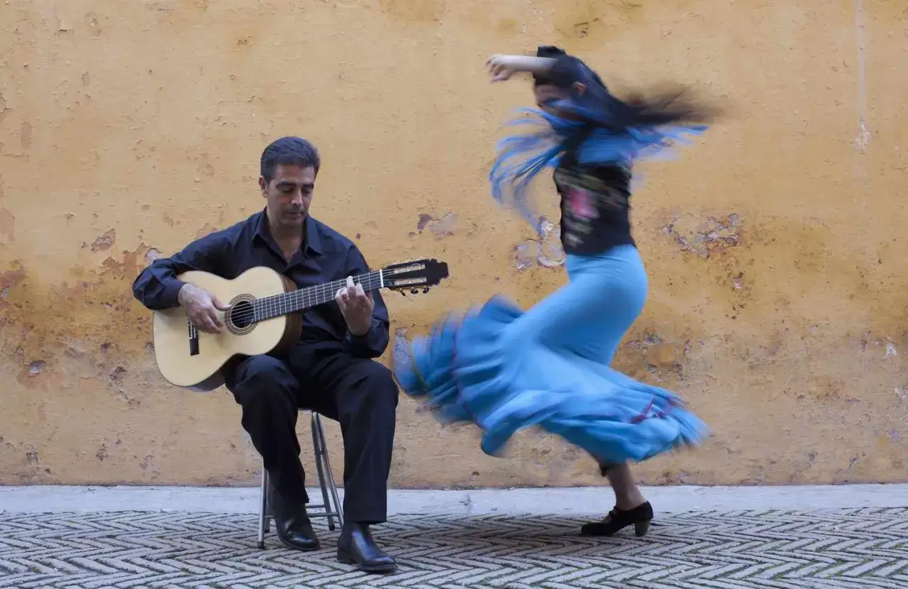 Hombre toca guitarra mientras bailarina con falda azul gira, capturando la esencia de de donde es la rumba.