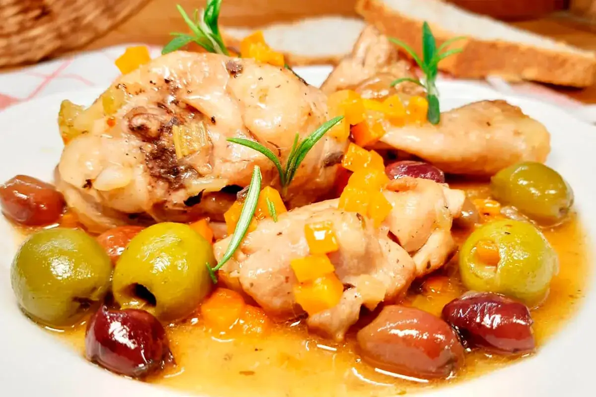 Un saporito pollo alla cacciatora con olive verdi e nere, carote a cubetti e rosmarino, servito su un piatto bianco.