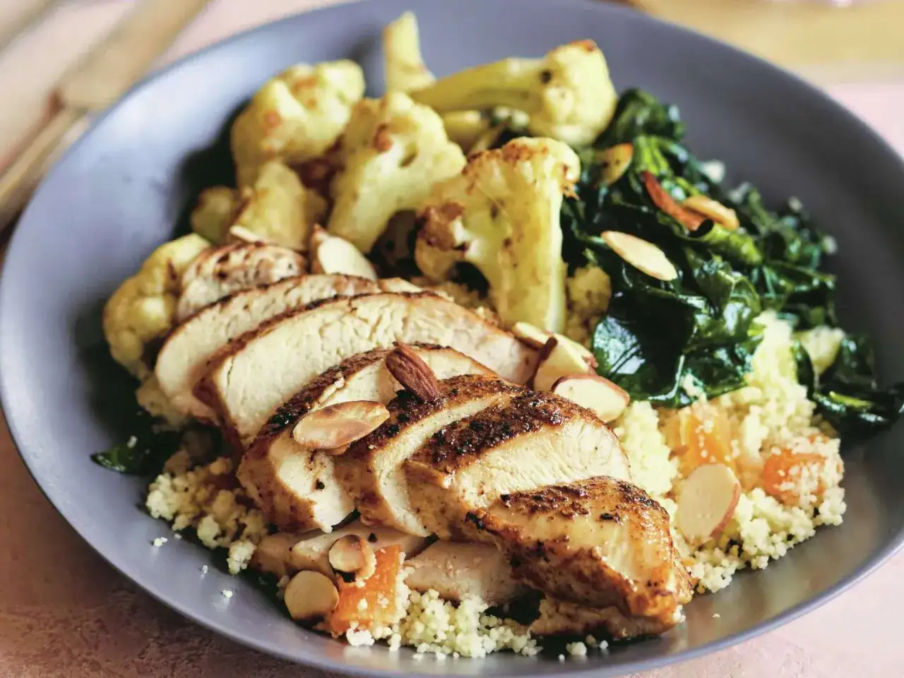 Piatto con cous cous, pollo grigliato a fette, cavolfiore arrosto e spinaci saltati. Un'idea su cosa abbinare al cous cous per un pasto completo e gustoso.