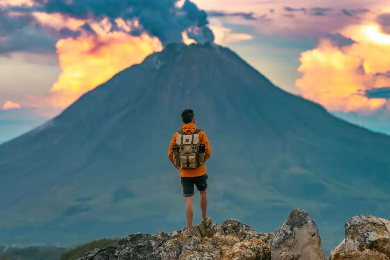 Ein Rucksacktourist steht auf einem Felsen und blickt auf einen rauchenden Vulkan. Ein unvergessliches Erlebnis beim Sumatra Backpacking.