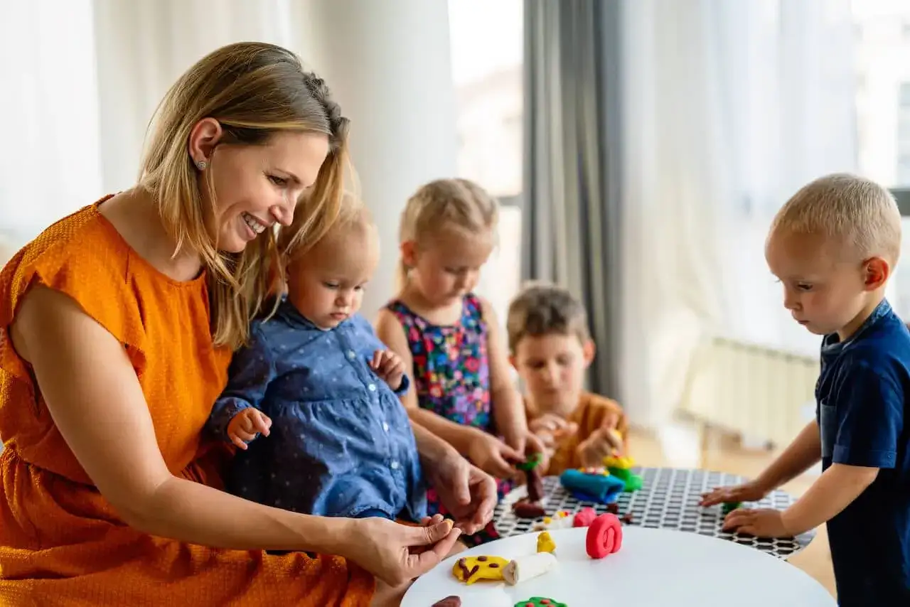 Frau spielt mit Kindern, die sich fragen, ab wann Kindergarten. Sie formen bunte Figuren aus Knete.