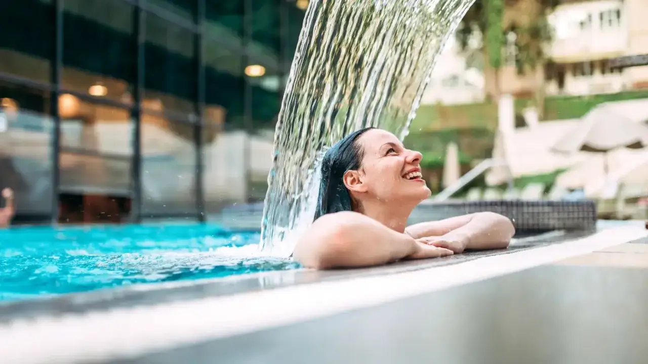 Mujer sonriendo bajo una cascada en una piscina, disfrutando de los balnearios de la Comunidad Valenciana.