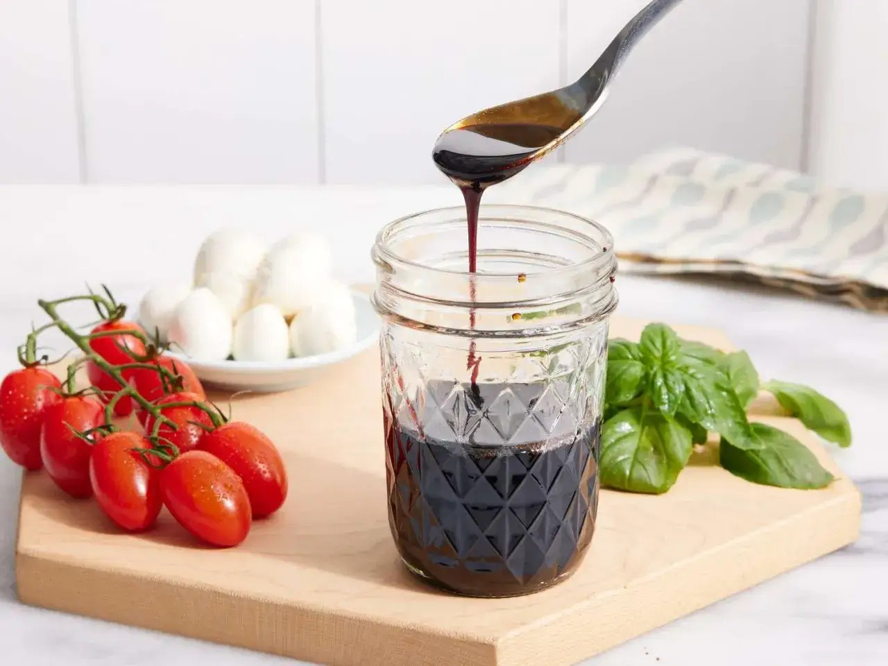 Gęsty, ciemny sos balsamiczny spływa z łyżki do słoika. Obok pomidorki koktajlowe, kulki mozzarelli i listki bazylii.