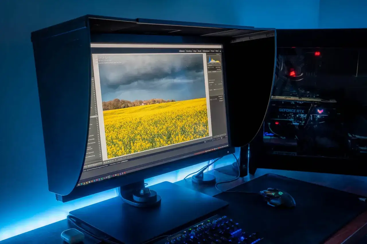 Profesjonalny monitor do obr&oacute;bki zdjęć z widokiem na pole rzepaku i burzowe niebo. Obok komputer z podświetlaną kartą graficzną.