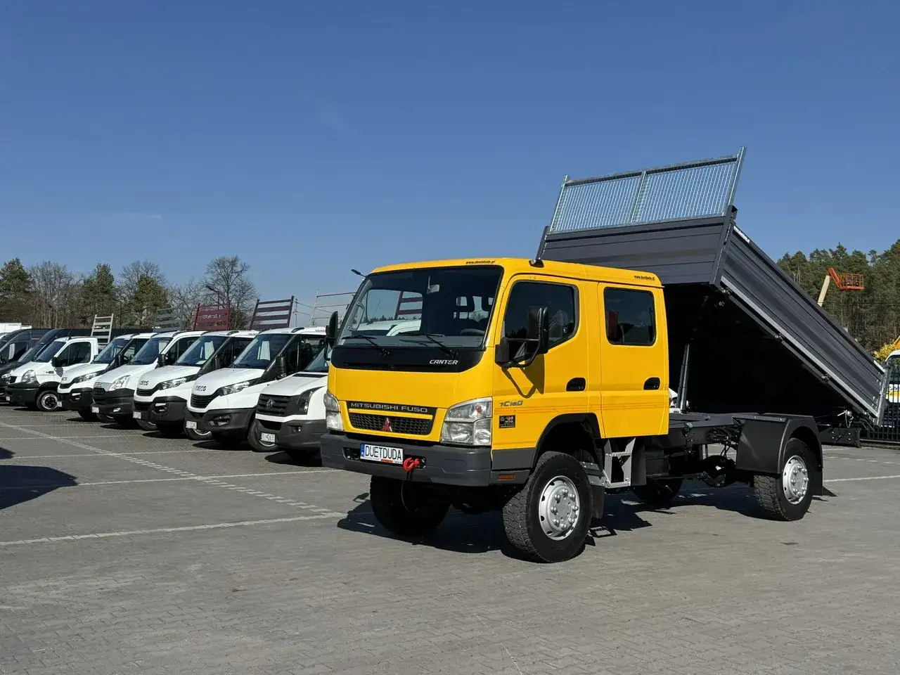 Żółty wywrotka Mitsubishi Canter z podniesioną skrzynią ładunkową stoi na parkingu. W tle rząd białych vanów.