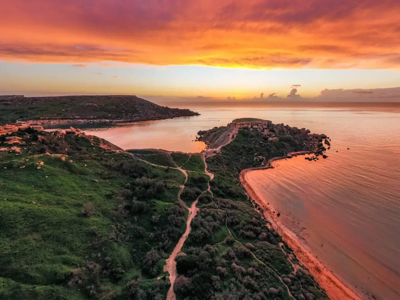 Zachód słońca nad Riviera Beach Malta. Pomarańczowe niebo odbija się w spokojnym morzu, a zielone wzgórza schodzą ku piaszczystej plaży.