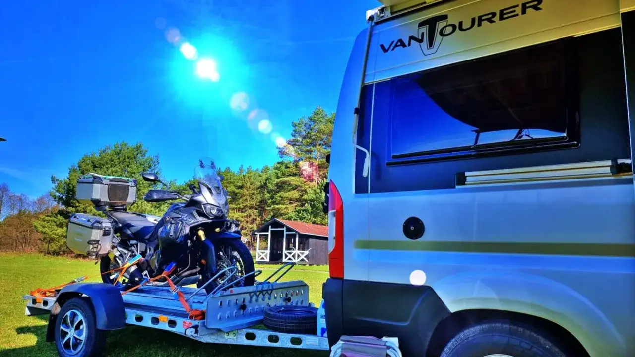 Motocykl na przyczepce obok kampera, gotowy na wyprawę po Sudetach i Karpatach. Słoneczny dzień zapowiada piękne widoki.
