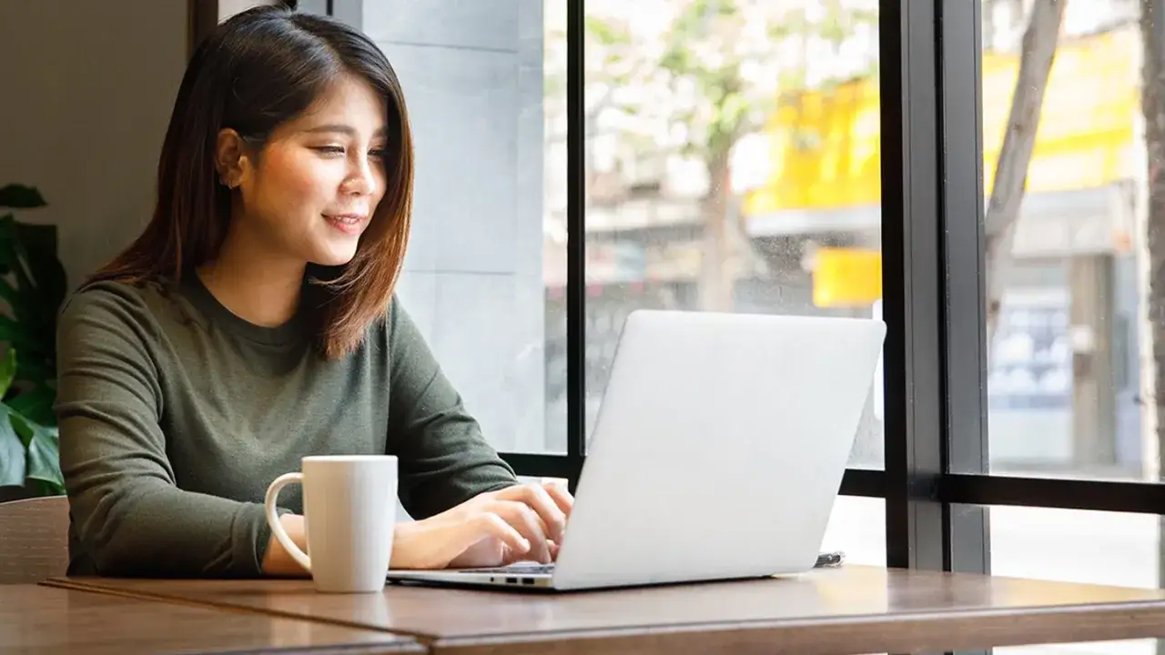 A young woman smiles while typing on a laptop, researching online MPA programs no GRE. A coffee mug sits beside her.