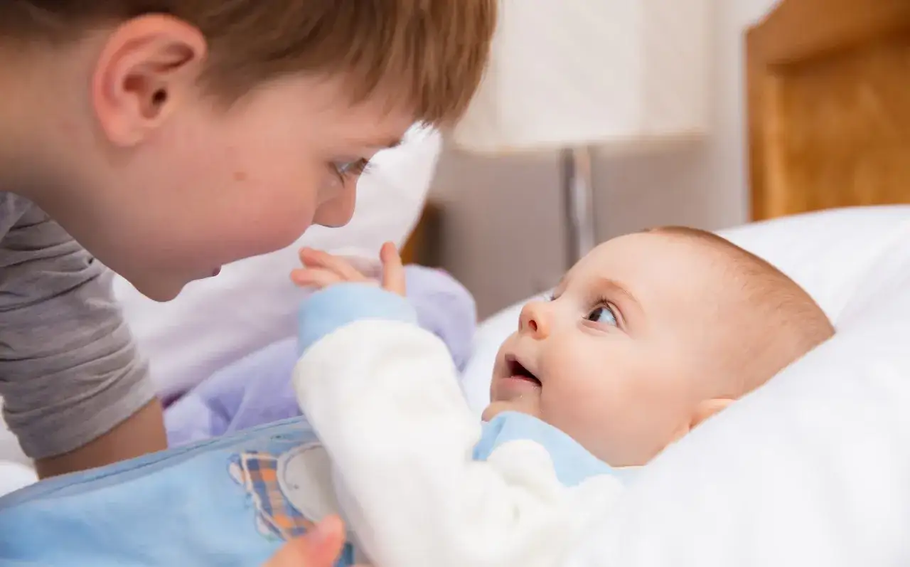 Starszy brat pochyla się nad niemowlakiem, który leży na poduszce. Dzieci patrzą na siebie z czułością, mimo że maluch ma objawy refluksu.