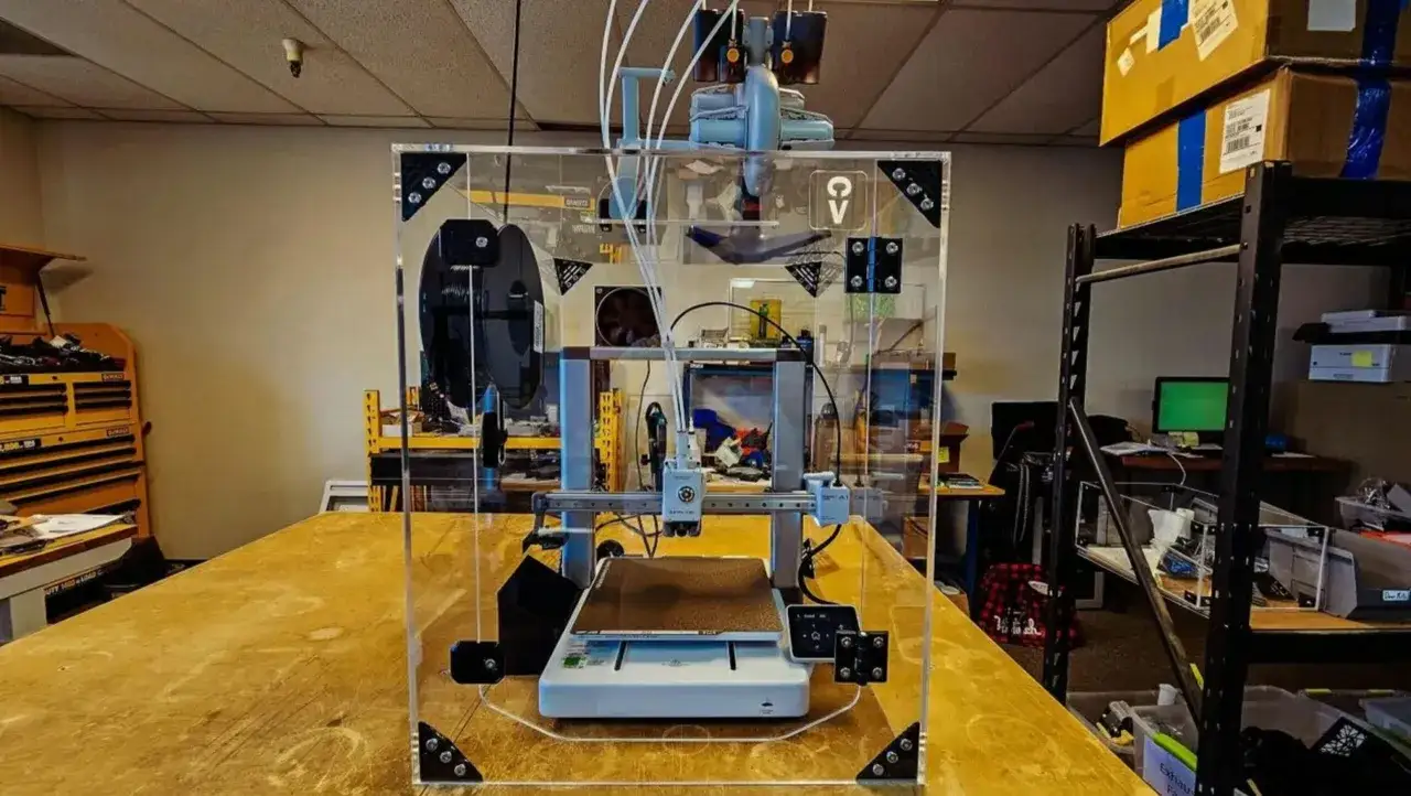 Drukarka 3D Bambu Lab w akrylowej obudowie, gotowa do pracy. W tle warsztat z narzędziami i regałami.