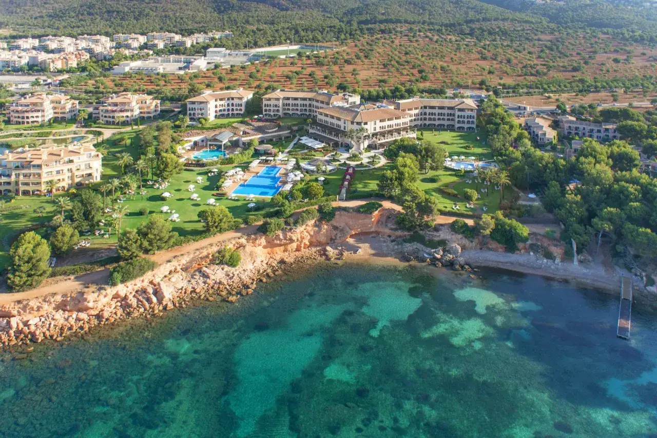 Luftaufnahme des Grupotel Natura Playa mit mehreren Gebäuden, Pools und gepflegten Grünflächen, die an eine felsige Küste und das türkisfarbene Meer grenzen.