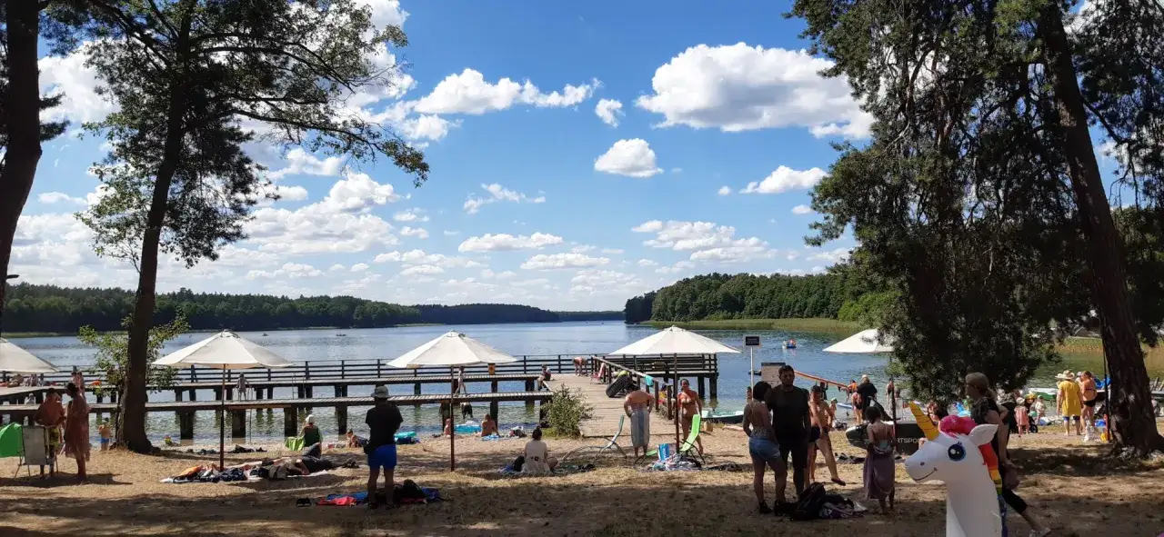 Słoneczny dzień nad jeziorem. Ludzie odpoczywają na plaży i spacerują po pomoście. W tle las i błękitne niebo z chmurami.