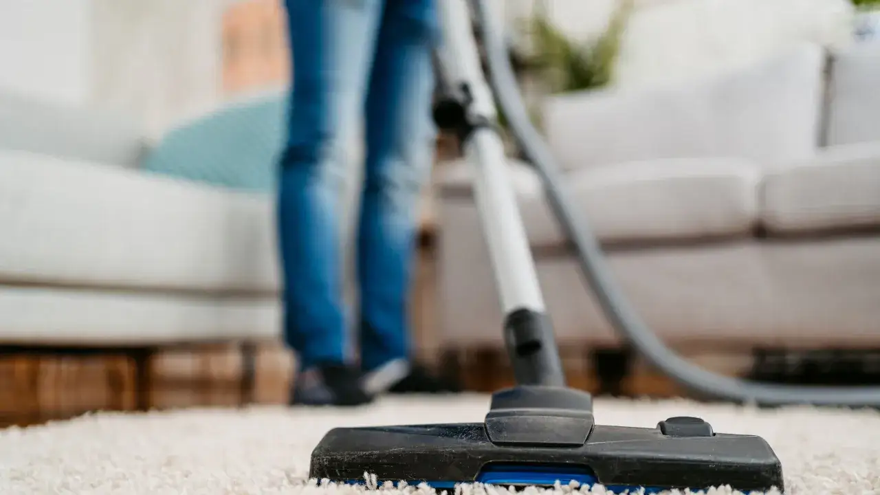 Person saugt Teppich trocken reinigen. Der Staubsaugerkopf ist nah am hellen Teppich, im Hintergrund sind Beine in Jeans und ein Sofa zu sehen.