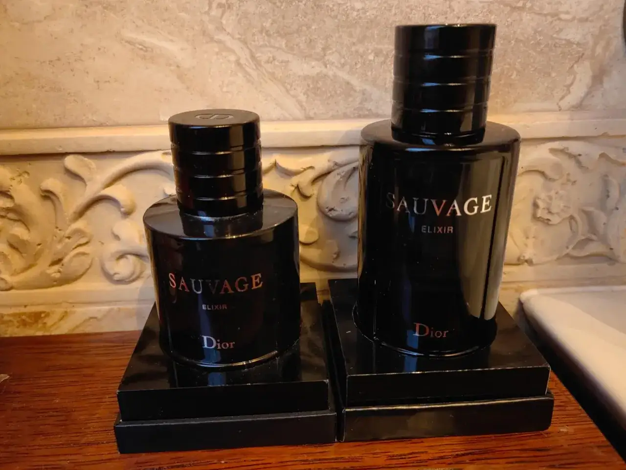Dwa eleganckie flakony perfum męskich Dior Sauvage Elixir, stojące na czarnych podstawkach, emanują luksusem.