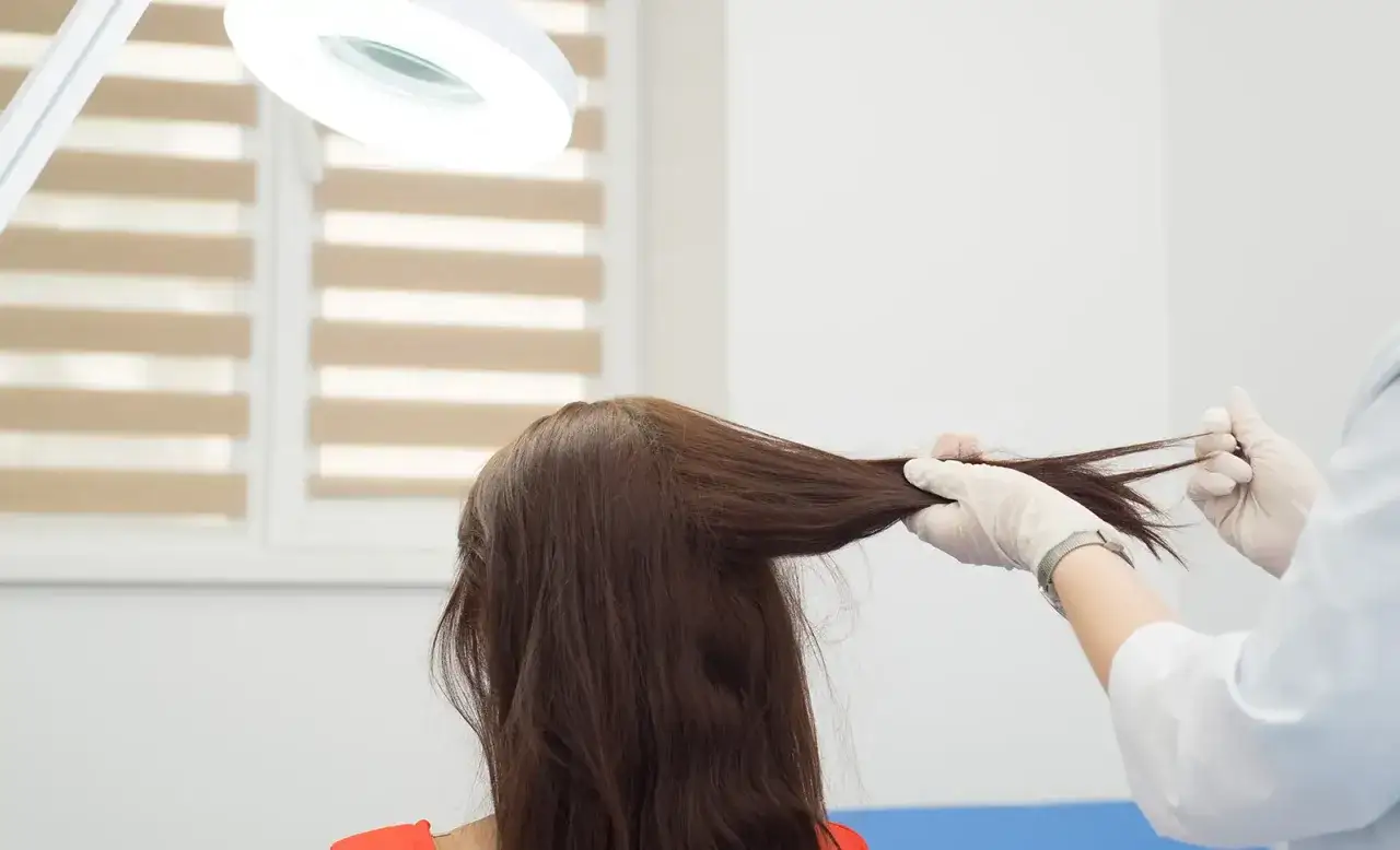 Mani guantate eseguono un **pull test** sui capelli di una donna sotto una lampada ingrandente, valutando la salute del cuoio capelluto.