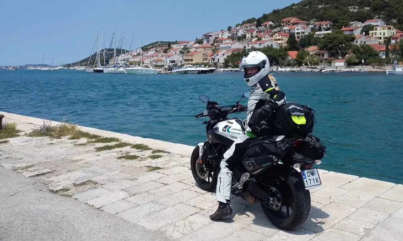Motocyklista w pełnym stroju na tle malowniczego wybrzeża z łodziami. To jak podróż po **autostradzie sudeckiej**, tylko że nad morzem!