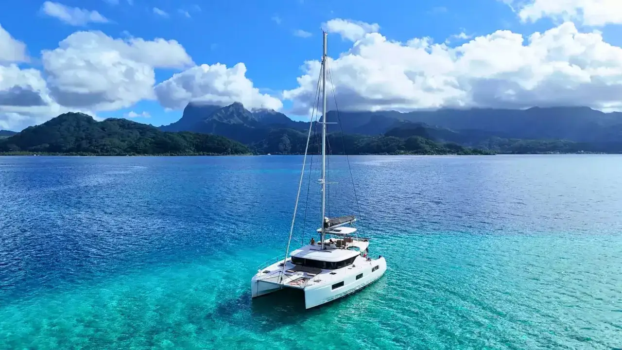 Un catamaran blanc, prêt pour un voilier tour du monde, navigue sur des eaux turquoise cristallines, avec des montagnes verdoyantes en arrière-plan.