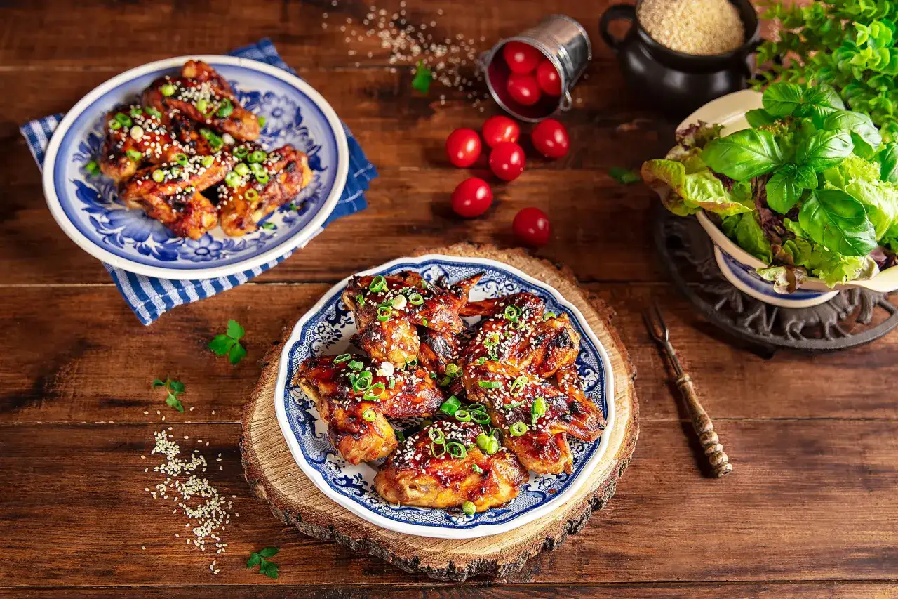 Pyszne skrzydełka teriyaki, błyszczące od sosu, posypane sezamem i szczypiorkiem, na talerzu z niebieskim wzorem.