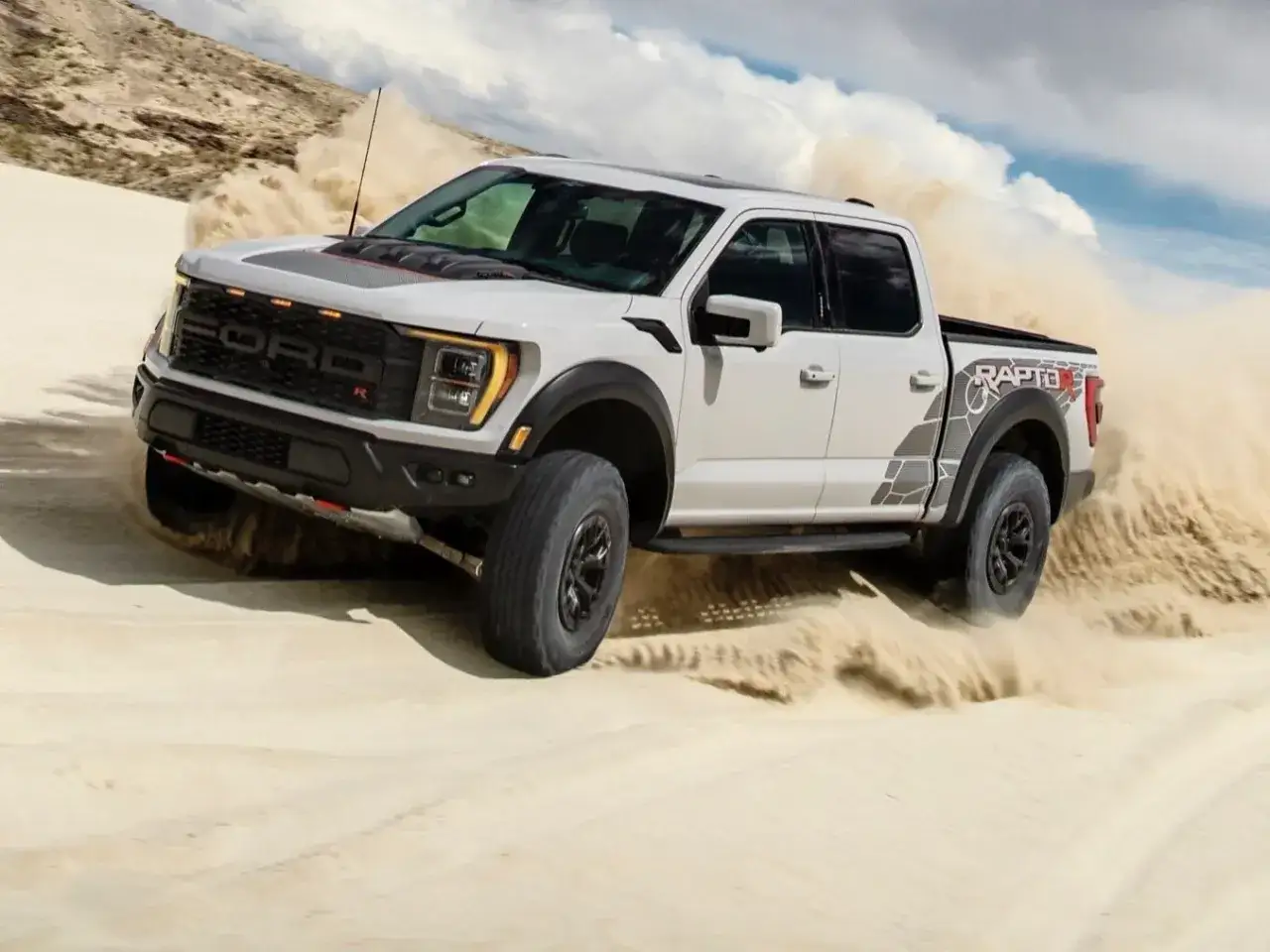 Biały Ford F-150 Raptor R szaleje po piaszczystych wydmach, wzbijając tumany piasku.
