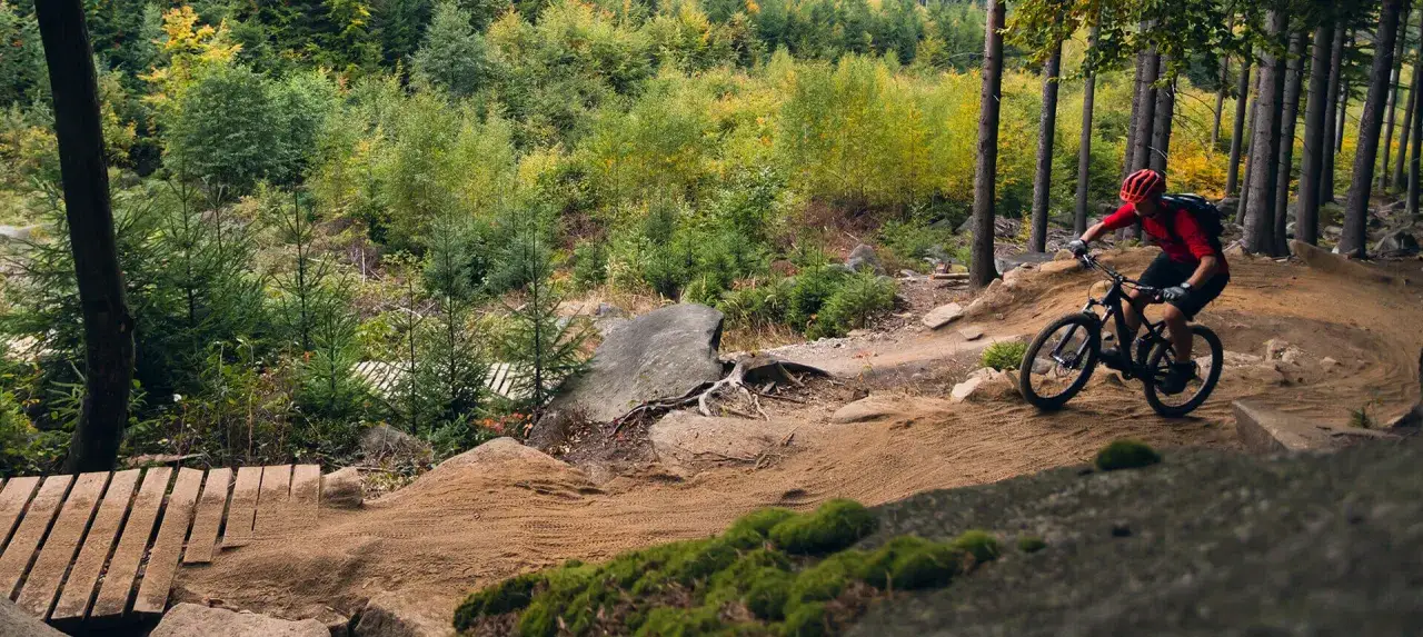 Rowerzysta w kasku pokonuje zakręt na piaszczystej, fortecznej trasie rowerowej wśród jesiennego lasu.