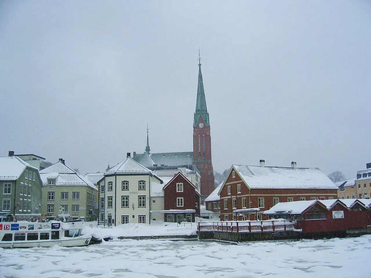 Vintervy &ouml;ver Arendal Norge med sn&ouml;kl&auml;dda tak och en kyrka med h&ouml;gt torn. Is p&aring; vattnet.