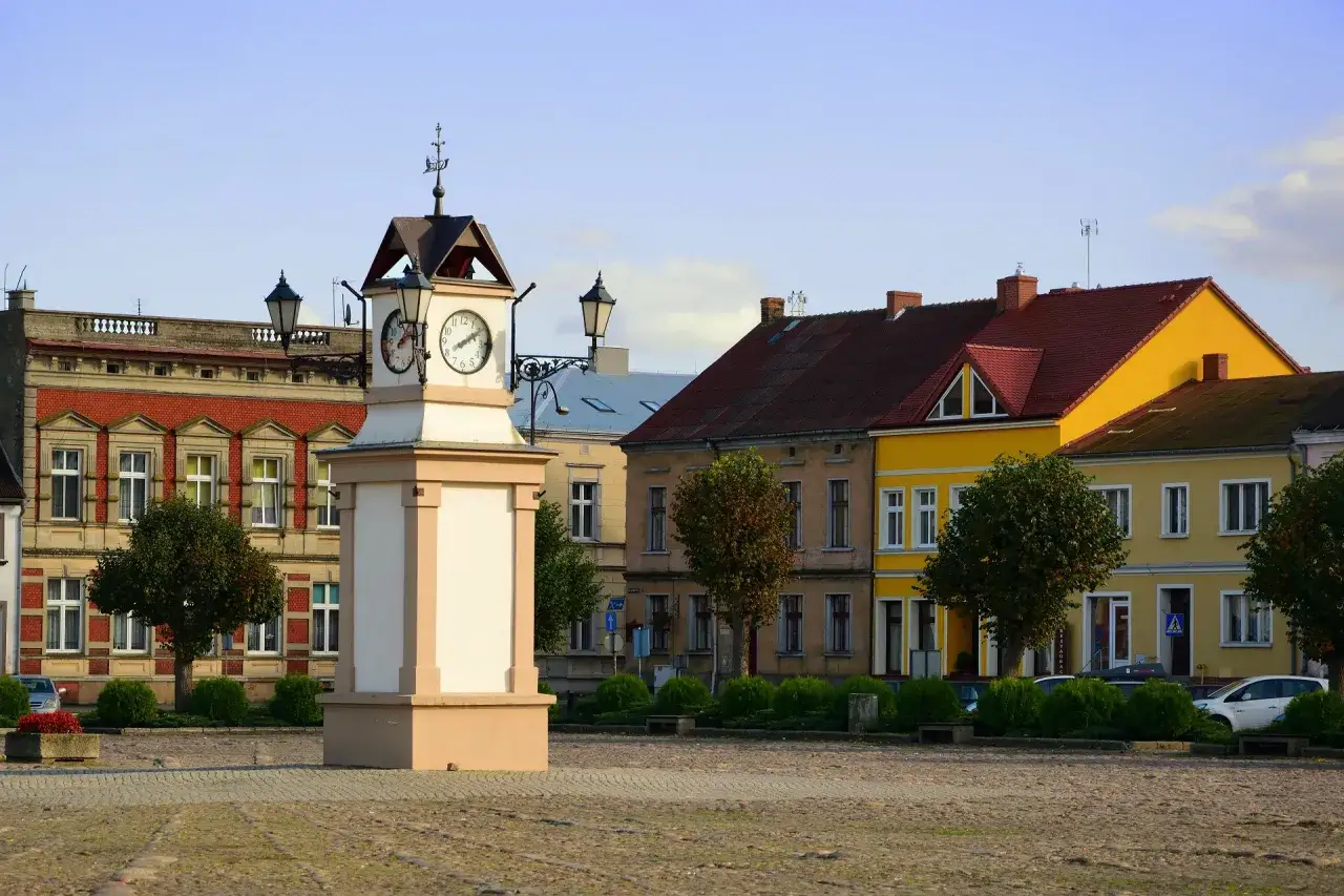 Rynek w **Lwówek Wlkp.** z zegarową wieżą po lewej i kolorowymi kamienicami w tle.