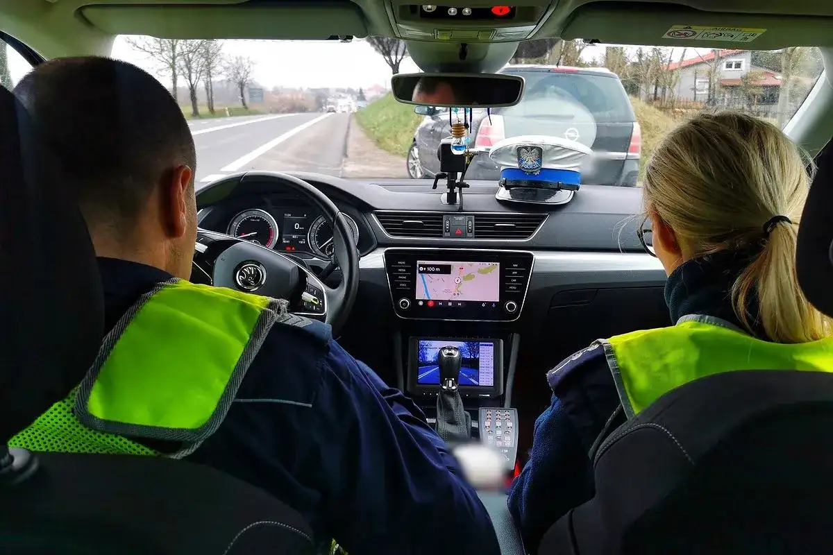 Policjanci w radiowozie, jadąc drogą, sprawdzają, czy trzeba wozić prawo jazdy. Na desce rozdzielczej widoczna czapka policyjna.