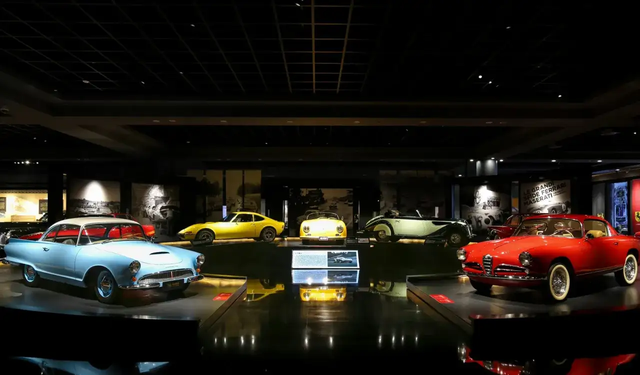 Kolekcja lśniących, **starych sportowych aut** w muzeum. Błękitny klasyk z białym dachem, żółty sportowiec i czerwona Alfa Romeo przyciągają wzrok.