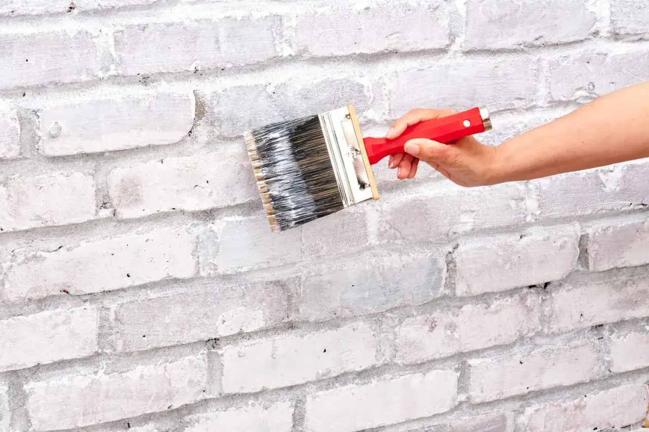 A hand applies white limewash to a brick wall with a wide brush, giving it a rustic, textured finish.