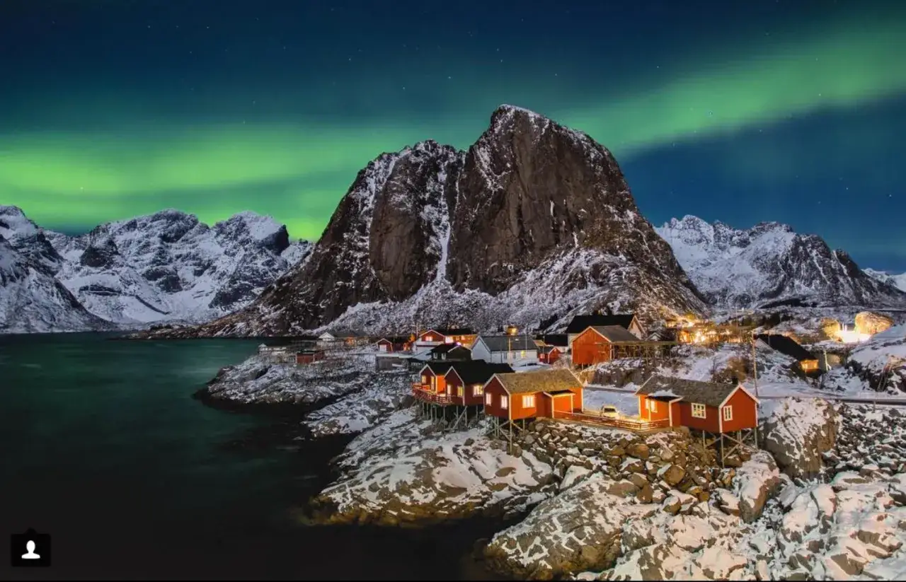 Vinter i Lofoten med norrsken som dansar &ouml;ver sn&ouml;kl&auml;dda berg och r&ouml;da fiskebodar vid vattnet.
